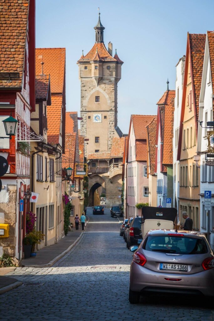 An upward view of a European cobblestone street showing red-tiled roofs, a central clock tower, and bright daylight.