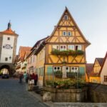 A picturesque European town featuring traditional half-timbered houses with steep roofs and a historic clock tower in the background.