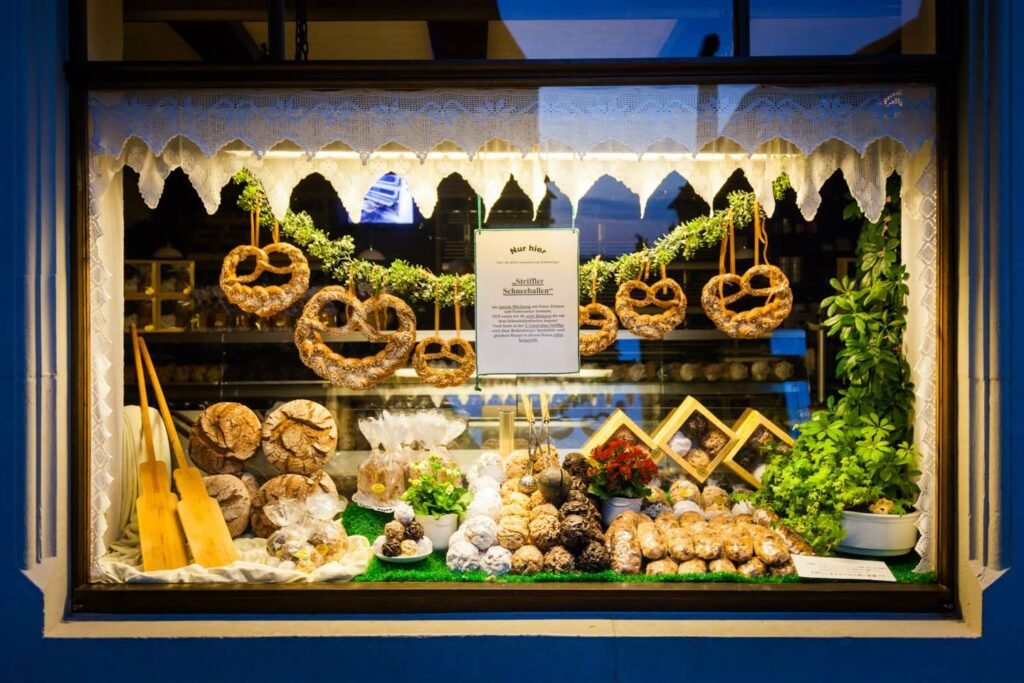 A festive bakery window display featuring pretzels, breads, and seasonal decorations. The storefront is brightly lit against a blue facade, showcasing an inviting arrangement of baked goods and plants.