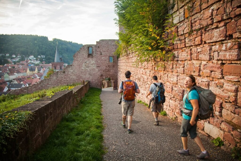 Three travelers with hiking backpacks walk along a stone path beside tall brick walls. A scenic view of a town and hills is visible in the distance.
