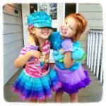 Two smiling children wearing matching prism dresses and a sequined cyan twill cap on a white porch.
