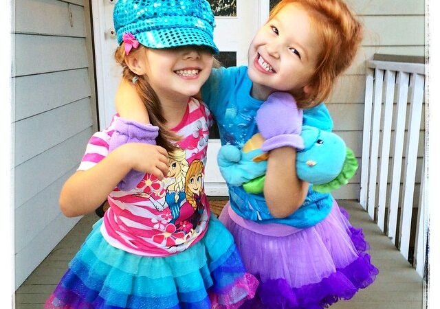 Two smiling children wearing matching prism dresses and a sequined cyan twill cap on a white porch.