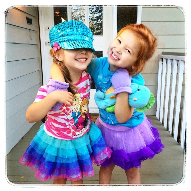 Two smiling children wearing matching prism dresses and a sequined cyan twill cap on a white porch.