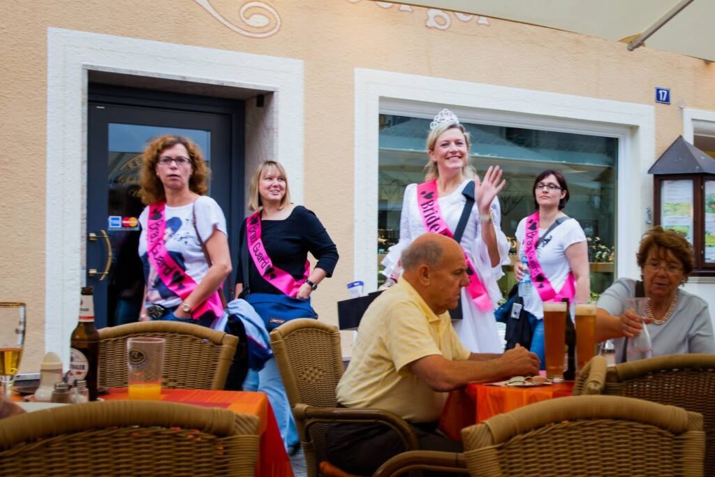 A group of women with pinks sashes and a person in a white headdress visit an outdoor café.
