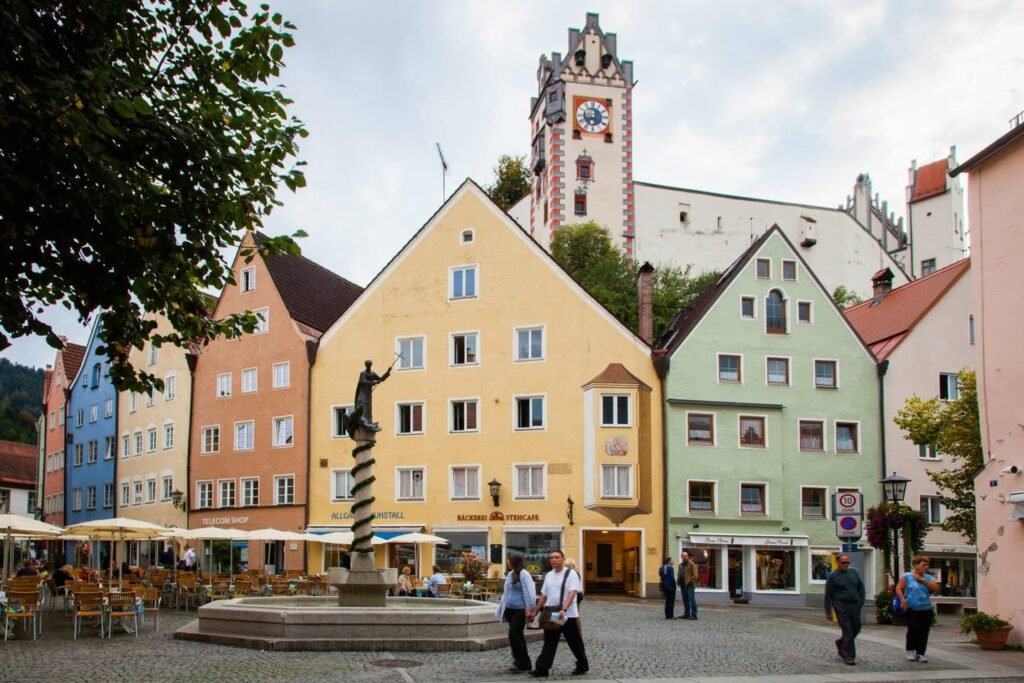 A European market square with brightly painted buildings and a castle tower in the background, where people are strolling and dining at outdoor tables.