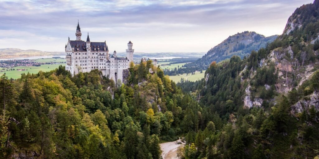 A majestic white castle with multiple turrets and spires perched on a forested hillside, overlooking a vast green valley.
