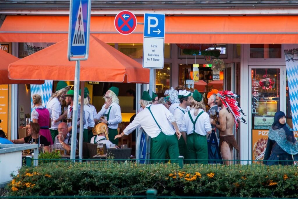 Men and women in traditional Bavarian attire at an outdoor Oktoberfest gathering, with orange umbrellas and beer signs visible.