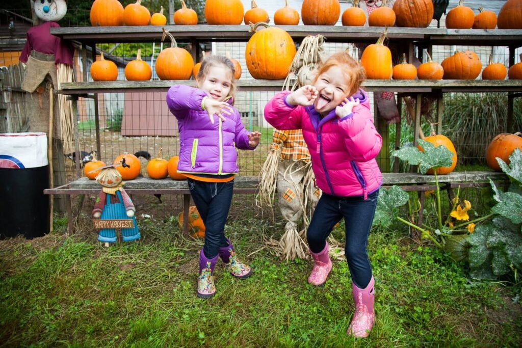 Two children in colorful jackets and boots enjoy a day at a local pumpkin patch. They play in the grass while orange pumpkins are displayed on wooden racks behind them.