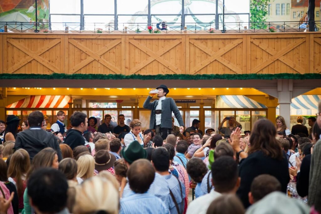 A vibrant crowd gathers at a wooden food venue, possibly Pike Place Market, for the Spring Storm food event. The lively setting features a person on stage addressing the audience amidst seasonal holiday decorations.