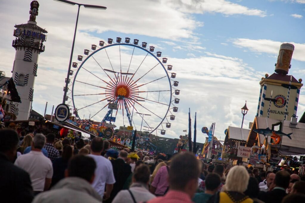 A bustling crowd gathers at the Downtown Lighthouse Fun Fair, with a prominent Ferris wheel and ride towers against a bright sky.