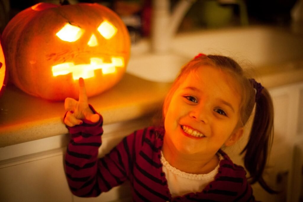 A smiling child in a striped shirt points toward a glowing carved pumpkin at night