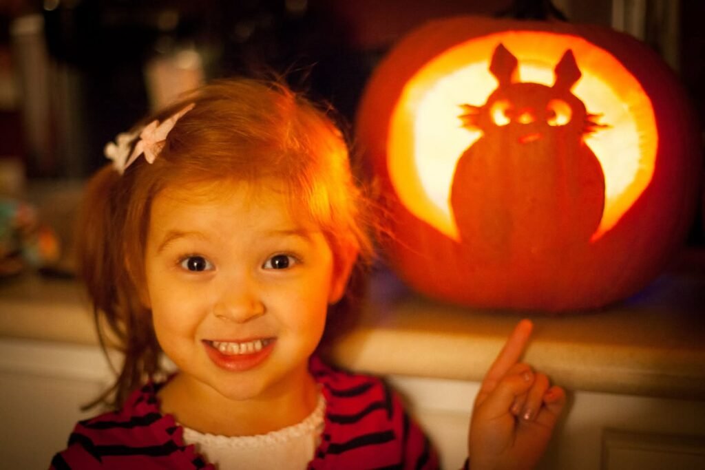 A smiling girl proudly posing with her owl-shaped carved pumpkin. A warm, glowing Halloween scene.