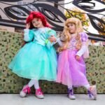 Two young girls dressed as princesses in teal and pink costumes pose in front of a mosaic wall.