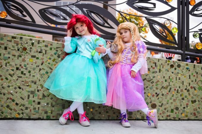 Two young girls dressed as princesses in teal and pink costumes pose in front of a mosaic wall.