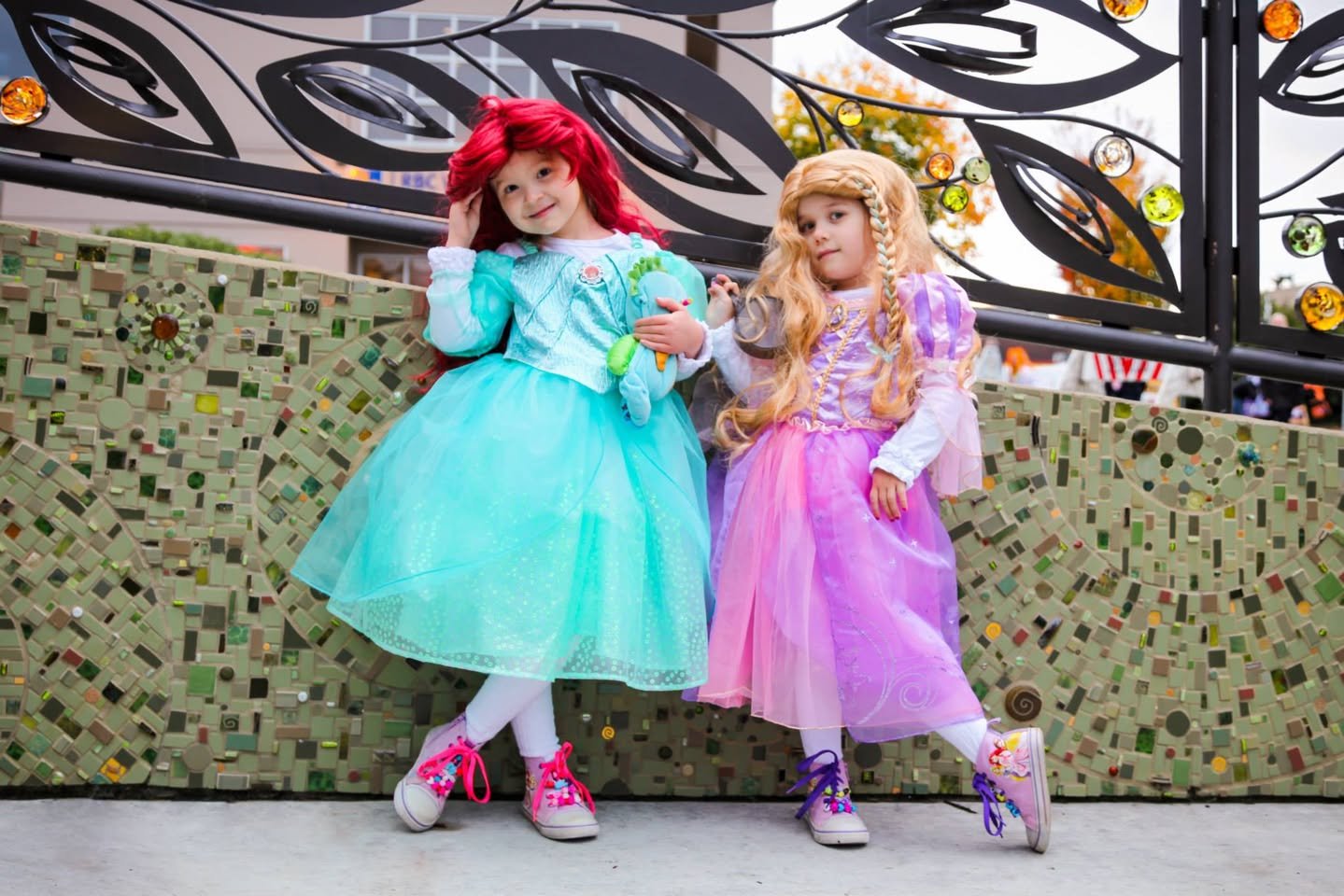 Two young girls dressed as princesses in teal and pink costumes pose in front of a mosaic wall.