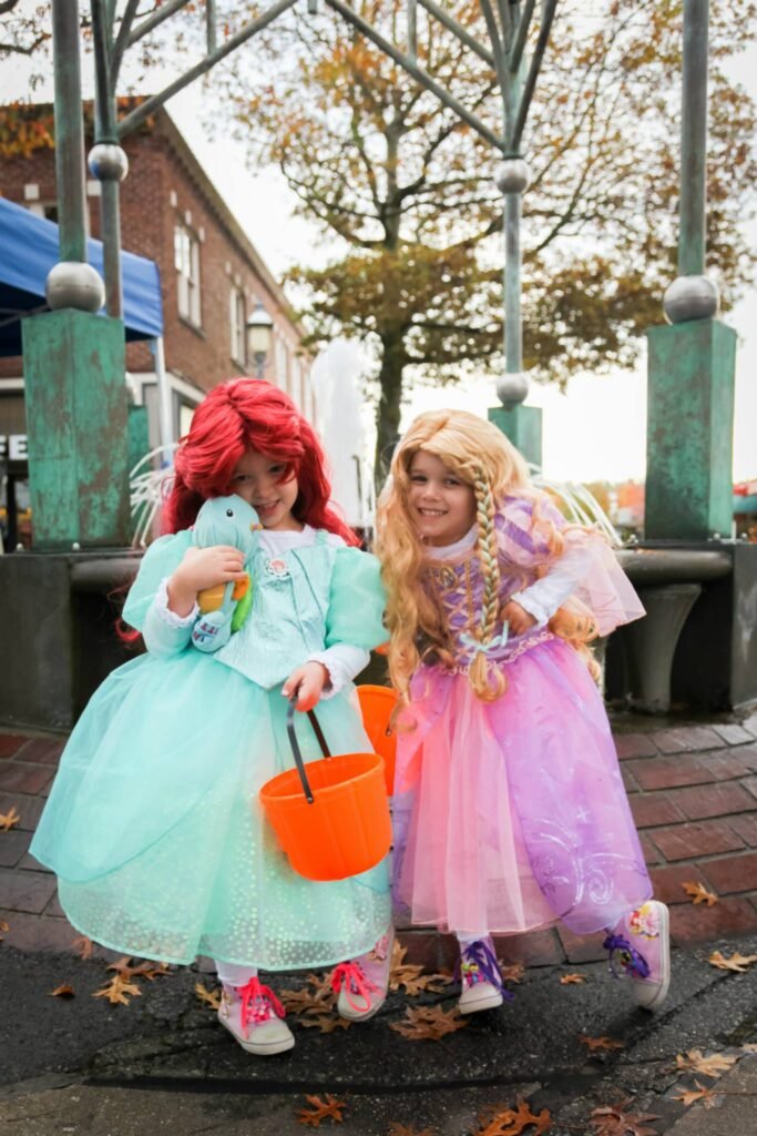 Two children in colorful princess costumes walk a brick sidewalk outdoors.