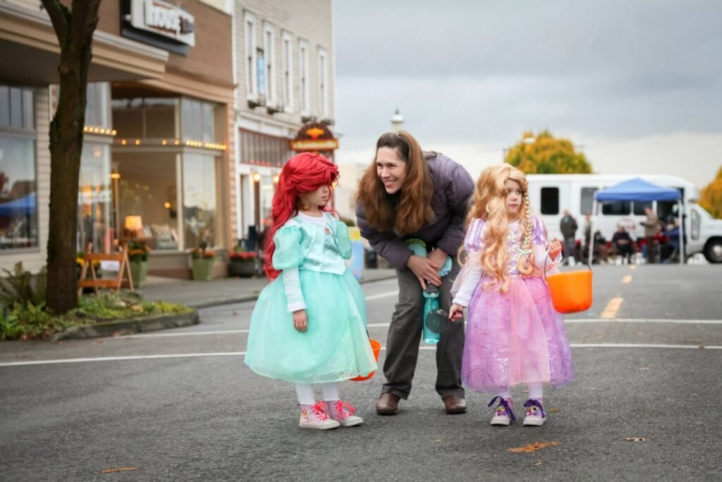 A long-haired woman crouched between two children in colorful princess costumes on a paved street in a small town.