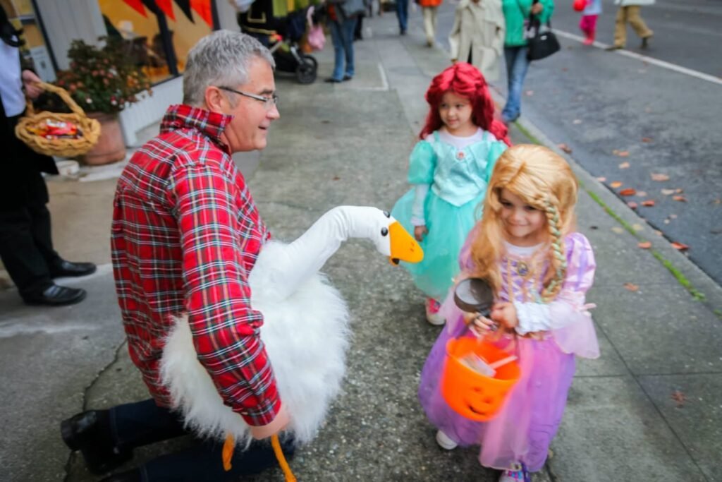 A person in a red plaid shirt and plush goose costume interacts with children dressed as princesses during an outdoor parade. The scene captures a festive moment on a public sidewalk.