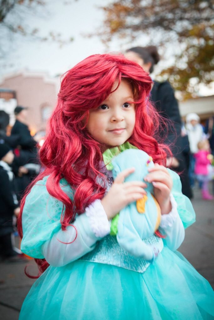 A young pirate princess wearing a red wig and holding a light blue plush toy at an outdoor event.