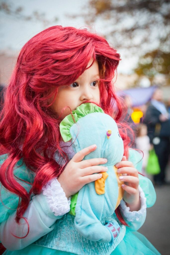 A child wearing a red wig and teal costume holding a light blue stuffed animal at an outdoor event.