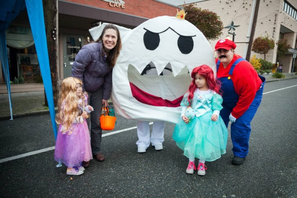 A smiling group wearing a purple dress, Mario outfit, and princess gowns poses beside a giant white ghost character on a city street.