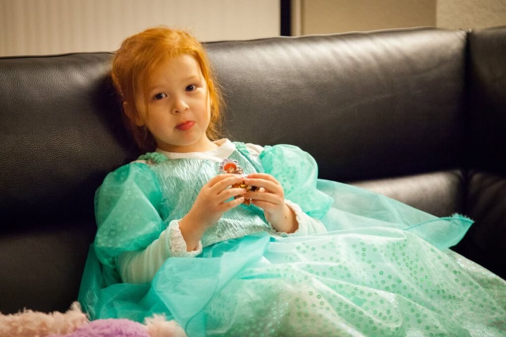 A young person with red hair wearing a light mint green princess-style dress while seated on a black leather couch. The subject holds a small object with both hands while looking at the camera.