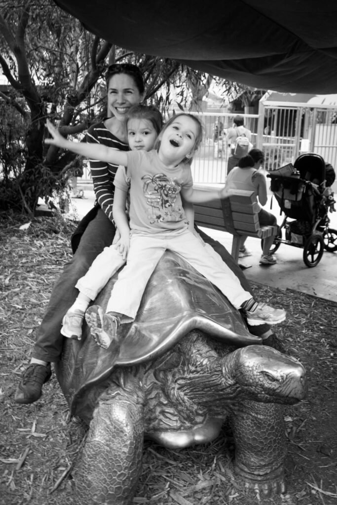 A joyful family poses on a large bronze turtle statue at a zoo, with a stroller and animal enclosure visible in the background. Close-up of the turtle's head and the kids' expressions.