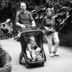 A black and white photo of a family walking a toddler in a stroller through a lush garden.