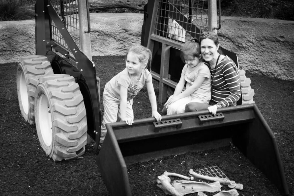 A black-and-white photo of three people sitting inside the bucket of a skid steer loader outdoors.