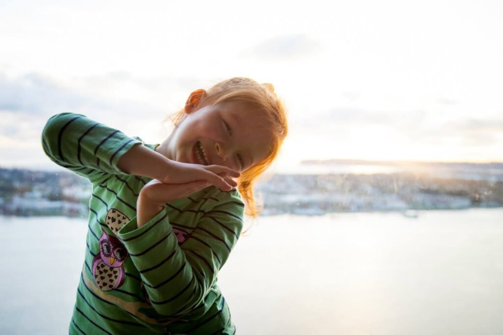 A happy child in a green striped sweater standing by a body of water during a bright, hazy sunset.