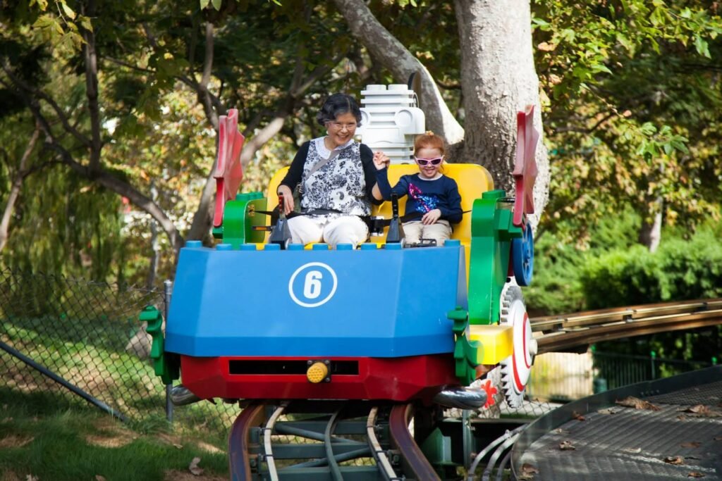 Two people riding on a colorful miniature train in an amusement park setting. The bright blue car marked with the number 6 travels along tracks.