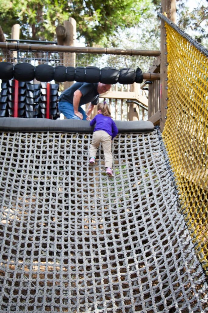 A view of a rope climbing wall in an outdoor play area with a child scaling the net and a padded handhold rail.