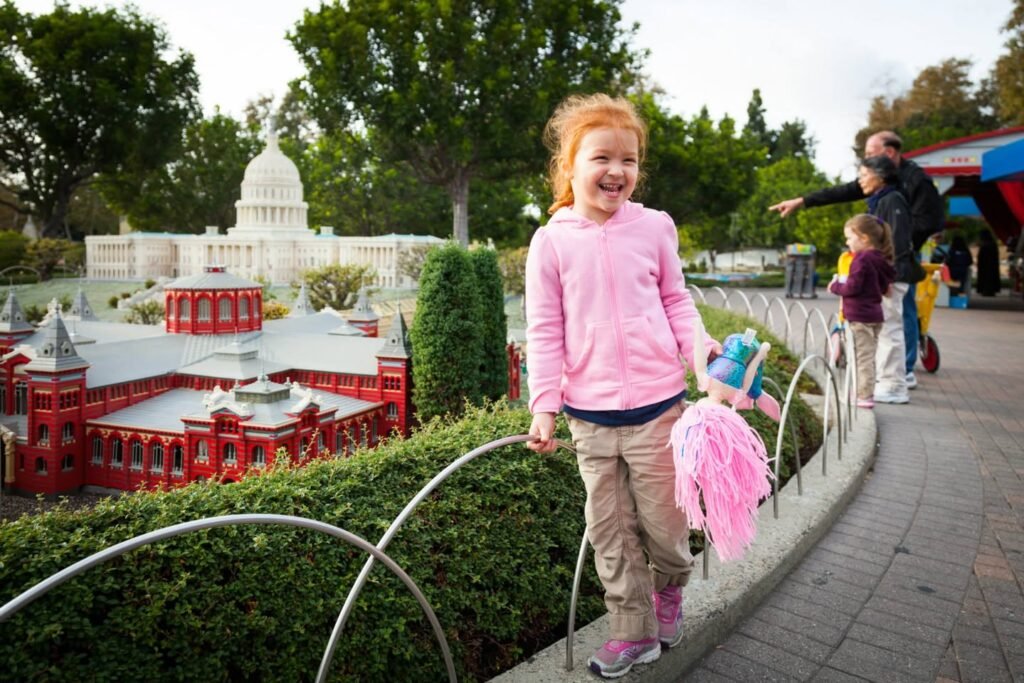 A child walks a paved path beside a miniature city model with a white domed building, while other visitors enjoy the display.