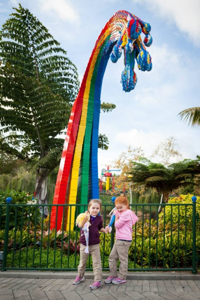 Two children stand in front of a large rainbow arch sculpture in a vibrant park. Lush palm trees and ornate green fencing add to the colorful scenery.