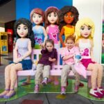 Children seated on a bench surrounded by large LEGO Friends figures.
