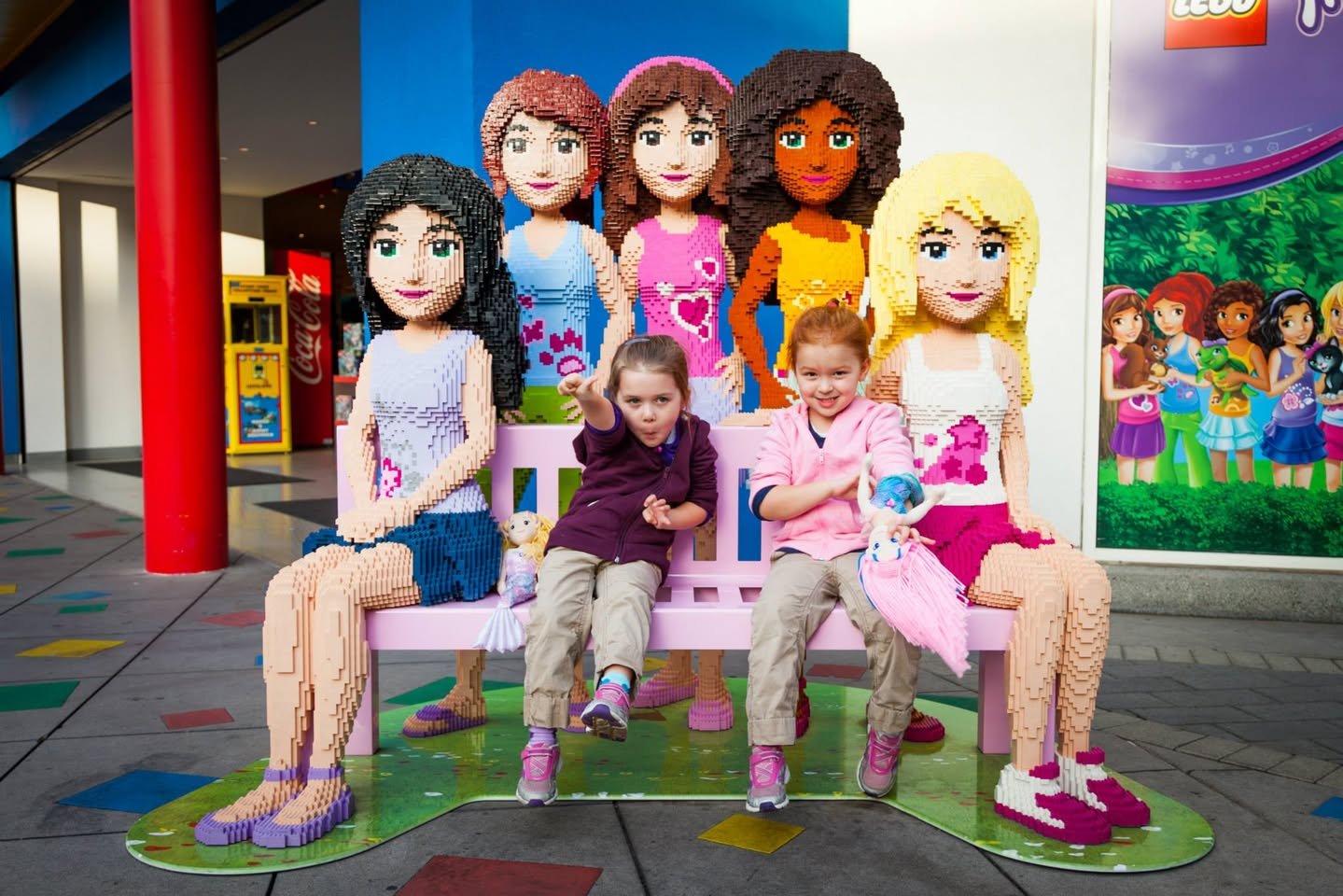 Children seated on a bench surrounded by large LEGO Friends figures.