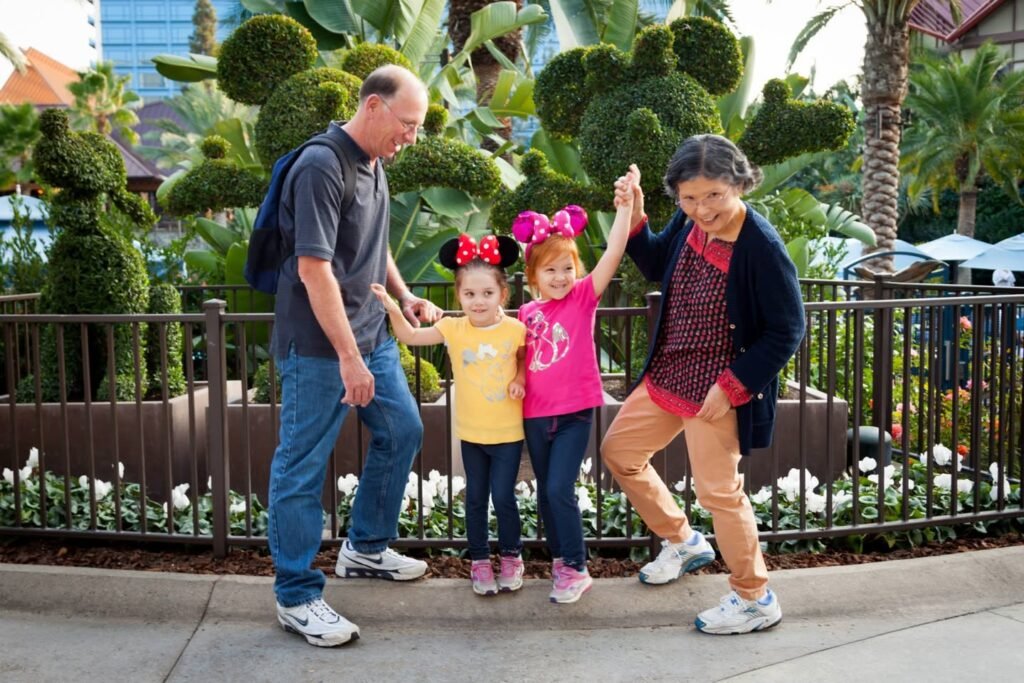 A family with two children wearing Minnie Mouse ears jumps joyfully in front of a topiary garden. White flowers and palm trees complete the scenic background.