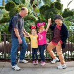 A family with two children wearing Minnie Mouse ears jumps joyfully in front of a topiary garden. White flowers and palm trees complete the scenic background.