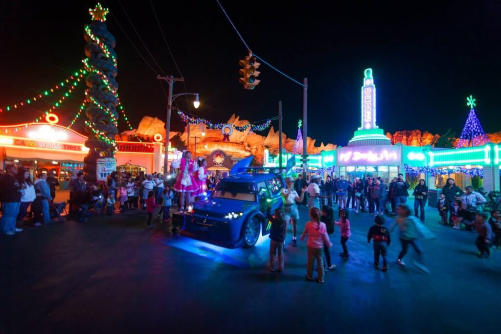 A nighttime holiday parade featuring illuminated floats, a decorated Christmas tree, and festive architecture.