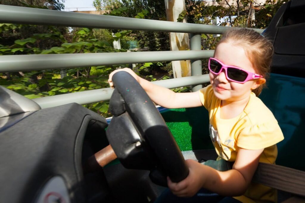A child wearing pink sunglasses smiles while holding the steering wheel of a kiddie ride with green foliage in the background.