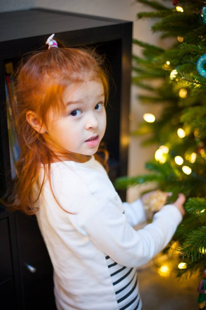 A young child in a white top decorates a green Christmas tree adorned with warm lights. The image captures a cozy holiday experience in a home setting.