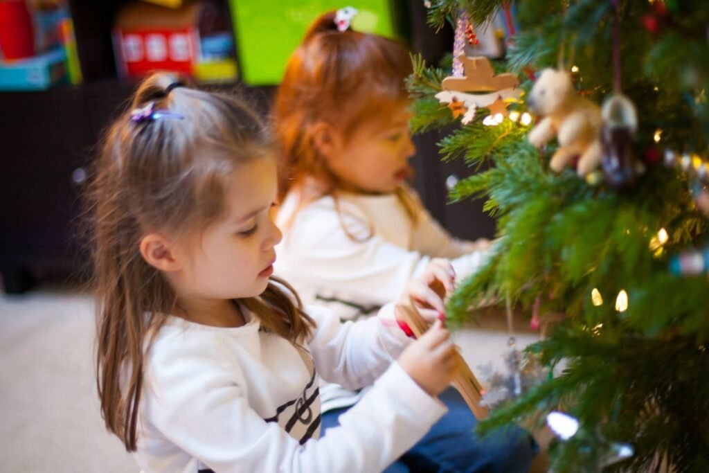 Two children in white shirts stand near a decorated Christmas tree with lights and ornaments.