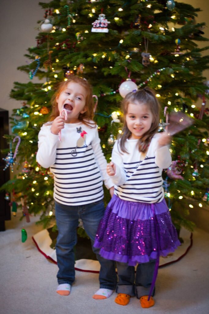 Two young children wearing striped shirts stand in front of a decorated Christmas tree with lights and ornaments.
