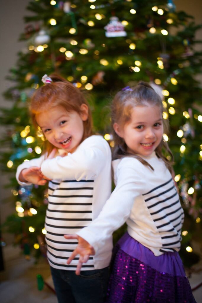 Two children posing back to back in striped shirts in front of a Christmas tree with warm lights.