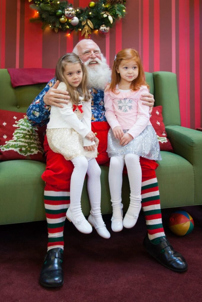 Children sitting on Santa's lap during a holiday visit. Festive decorations and green couch in the background.