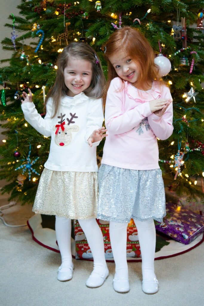 Two young girls wearing festive outfits and white tights stand smiling in front of a decorated Christmas tree.