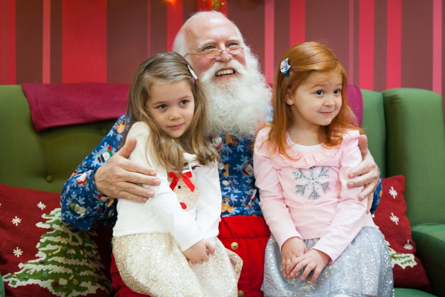 A man dressed as Santa Claus sits with two young girls on a green couch against a red striped wall. The scene features festive pillows and holiday cheer.