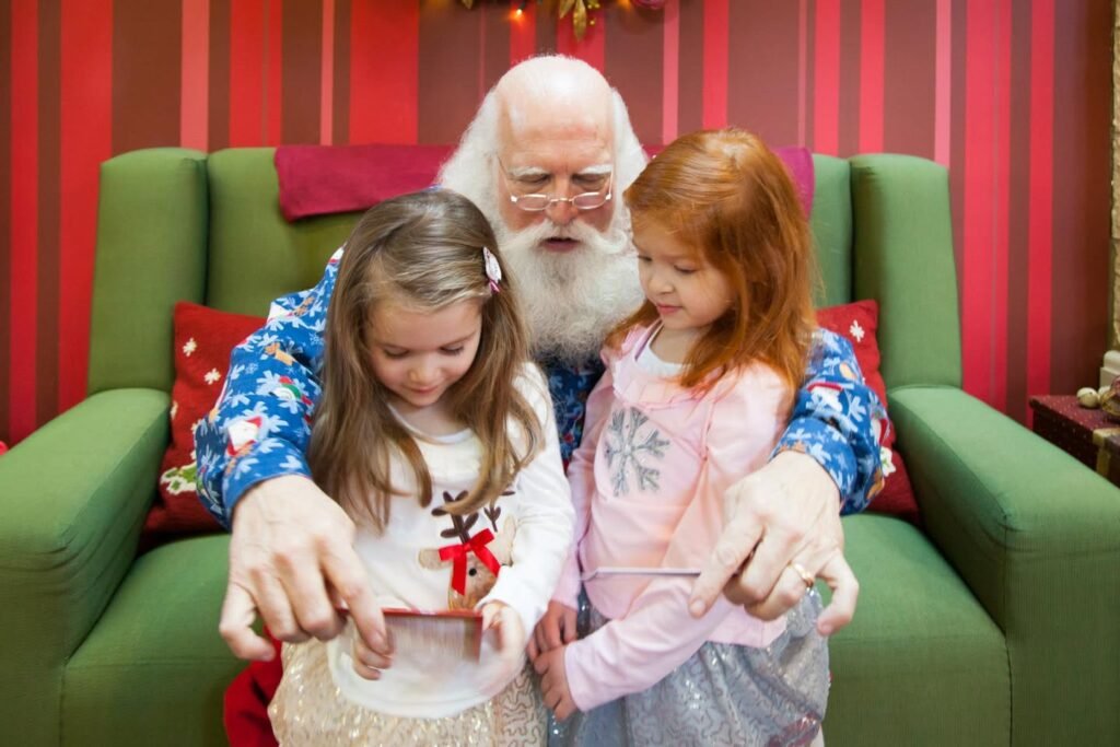 Santa Claus sits with two children on a green sofa. The group is focused on a book against a red-striped background.