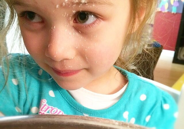 A child with flour on their forehead looks over a mixing bowl during baking.