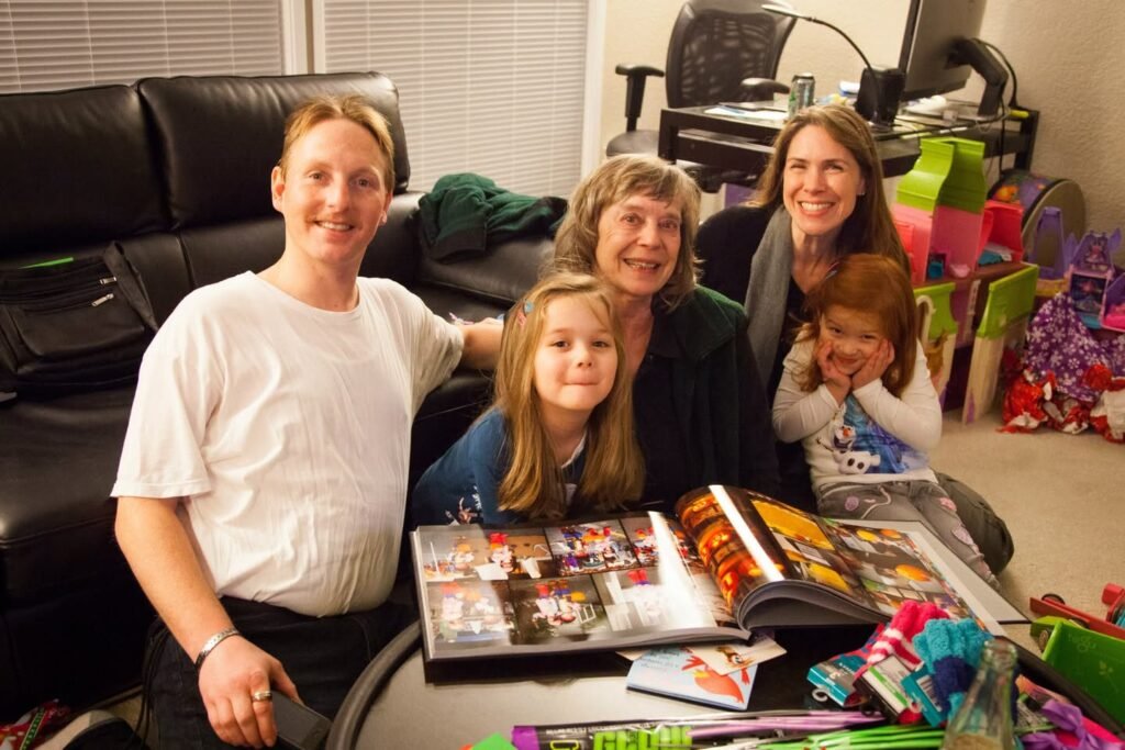 A group gathers in a living room to review photo albums. Someone holds a little one while colorful gifts sit nearby.